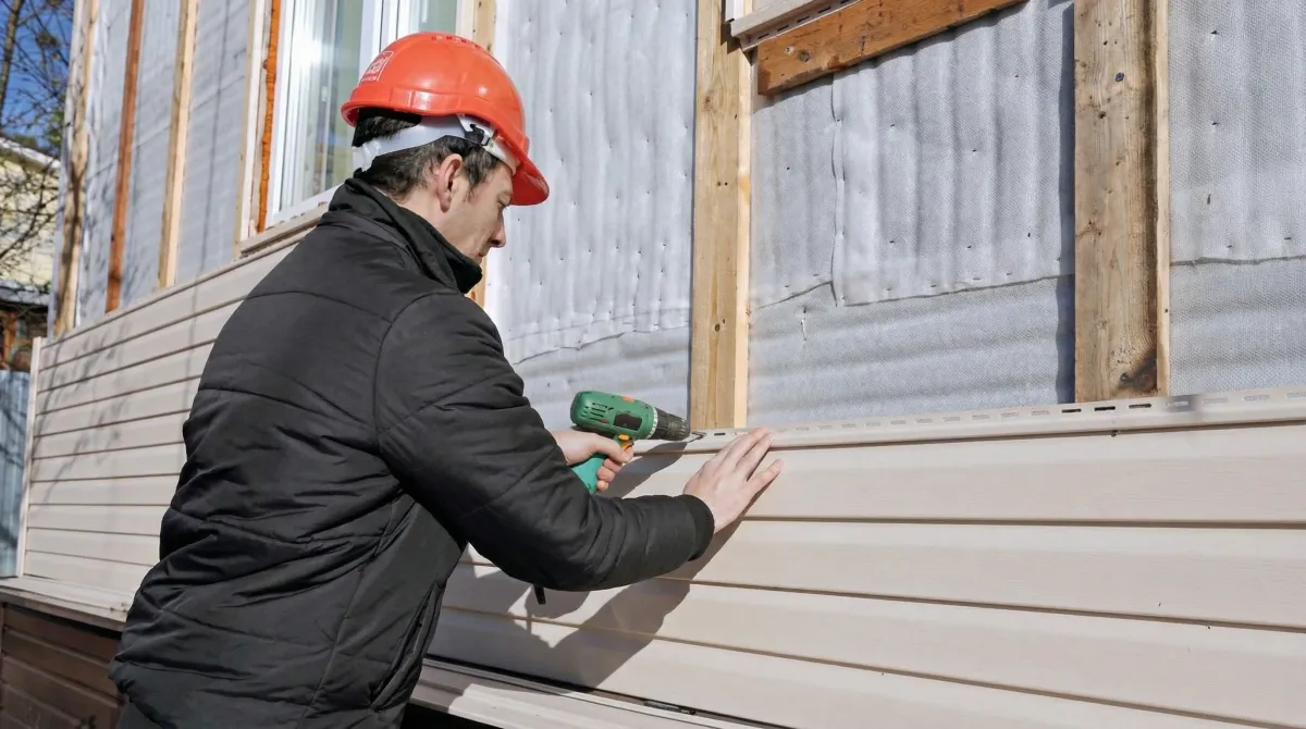 Siding replacement professional using a power drill to install new tan vinyl siding on a home exterior in Detroit, Mi